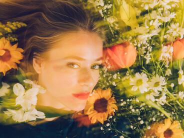 A women among flowers gazing up, surrounded by nature’s bloom, she lays in quiet confrontation