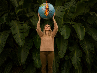 Beneath dense green foliage, a woman lifts a flaming Earth toward the sky, symbolizing humanity’s fragile relationship with nature and the growing urgency of the climate crisis