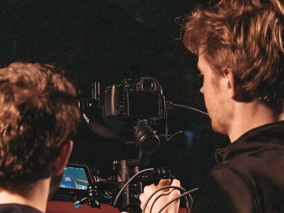 A man wearing a black shirt holds a black DSLR camera on a bustling Hollywood film set, surrounded by workers