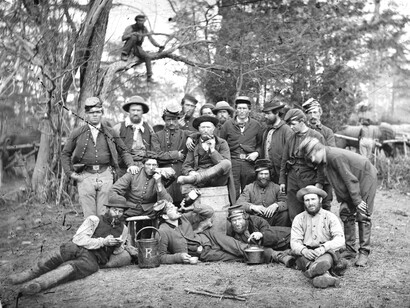 A May 1862 photo taken by James Gibson of a group of Union officers’ servants contains several peculiar details: one man drinks from a bottle, another seems to be reading a small book, a third clutches the handle of a kettle for unknown reasons, and in the background, one man has climbed onto a tree branch to pose for the camera