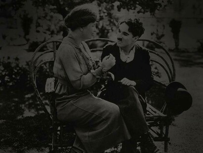 Helen Keller dressed elegantly is sitting with Charlie Chaplin in a park