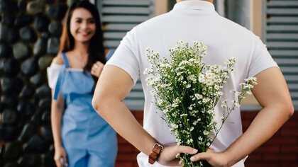 A man stands before a woman near a marble wall, tenderly presenting a bouquet of flowers, creating a scene of timeless romance