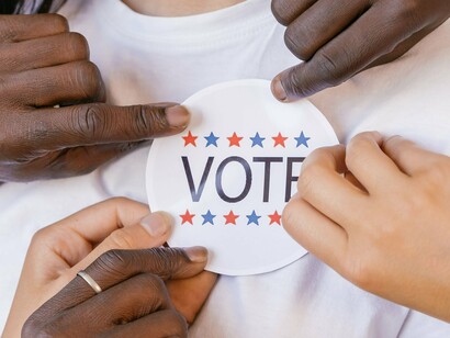 Mãos de diferentes pessoas seguram um adesivo relacionado ao voto diante de uma jovem sorridente. A democracia se constrói justamente pela participação de indivíduos diferentes em uma mesma esfera de escolha