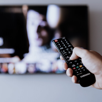 Una mano sostiene el mando mientras la televisión ocupa el fondo de la escena, en una imagen que recuerda la centralidad de la pantalla en el consumo cotidiano de información y entretenimiento