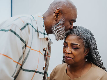 A senior man comforts his wife as she cries, holding her hand during her cancer treatment