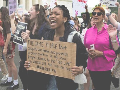 A black woman holding a cardboard sign that reads "The same people that support war, famine and poverty in other countries are the same people claiming to be 'pro-life' so whose life are they pro for?' 