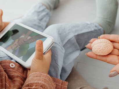 A child is captivated by a phone, while their parent sits nearby, holding a brain-shaped toy