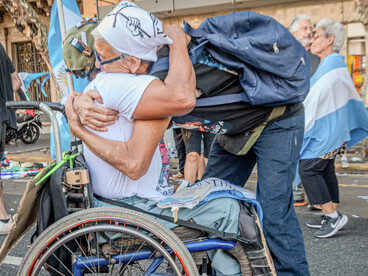 Momento emotivo durante una manifestación donde dos personas se abrazan cálidamente, una de ellas sentada en una silla de ruedas con banderas de Argentina de fondo