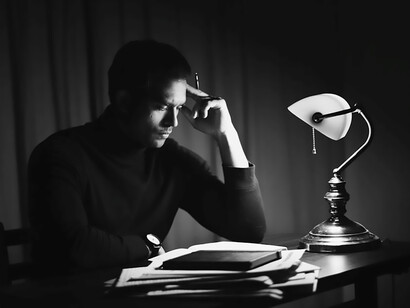 In a monochromatic room, a man wearing a black sweater is deep in contemplation as he pens his thoughts, engrossed in the process of writing a book