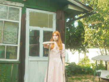 Bathed in warm summer light, a young woman in a vintage dress stands at the doorway of a rustic country house, framed by vibrant greenery and flowers
