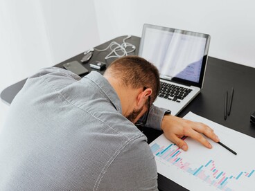Um homem de camisa cinzenta está debruçado sobre documentos, com a cabeça apoiada na mesa de trabalho. À sua frente, encontram-se um computador portátil e gráficos, enquanto algumas notas de dinheiro repousam sobre um caderno. A cena traduz o desgaste associado às pressões profissionais e financeiras, aproximando-se do alerta do autor sobre os custos de decisões equivocadas
