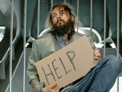 A homeless man sits on the ground, holding a banner asking for help