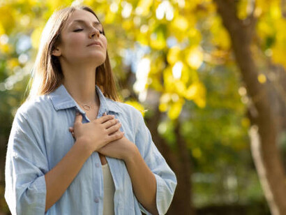 A woman gently places her hands on her chest, eyes closed, inhaling deeply in the tranquility of the forest