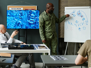 Military instructor pointing at a whiteboard covered with tactical sketches and directional arrows during a training session