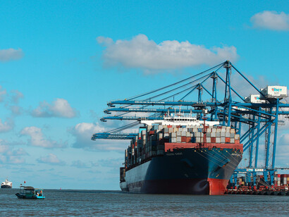 A cargo ship moves across the sea under a bright blue sky, approaching a busy harbor in China