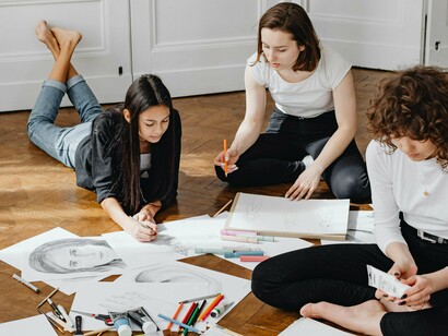 Três mulheres sentadas no chão, cercadas por folhas, desenhos e materiais de criação espalhados. Quando a ideia é apenas produzir, sem julgar, a criatividade encontra espaço para respirar