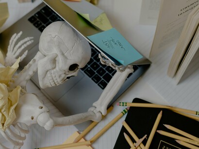 An overwhelmed skeleton buries its head in a laptop, surrounded by scattered books and pencils, reflecting the collapse of a work system where unsustainable demands turn effort into decay