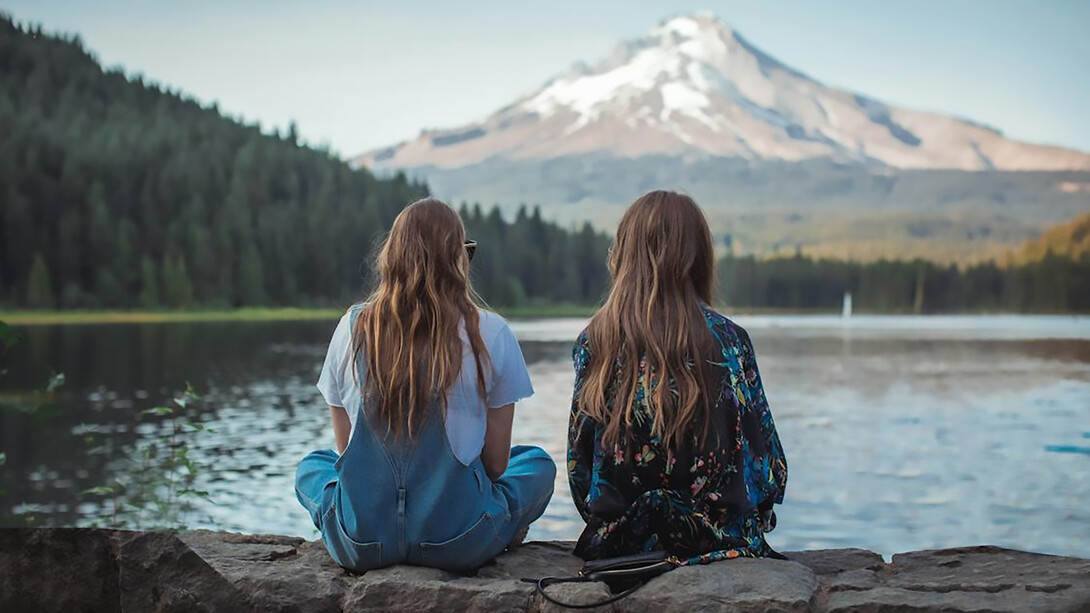 Two women sitting on the rock and looking at the water and mountain 