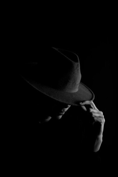 A man in a black suit wearing a white fedora, with the look of a classic crimina