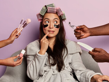 A young woman in a plush bathrobe applying under-eye patches, illustrating the impact of digital face-tuning and beauty industry consumerism