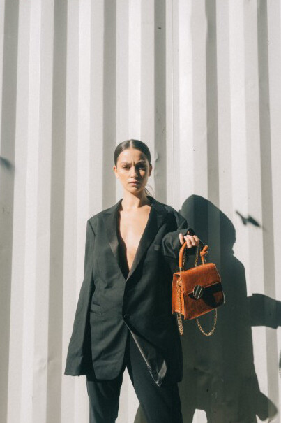 A woman in a black blazer, holding a brown handbag, leaning casually against a white wall, exuding old money fashion, generational wealth style, and timeless preppy elegance