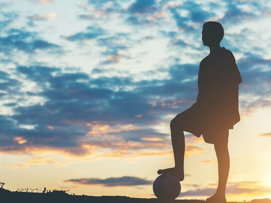 A young footballer silhouetted against the evening sky with a ball at their feet, representing dreams that reach far beyond the horizon