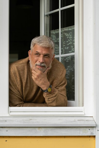Um homem inclina-se para fora da janela de uma casa amarela, observando o exterior. A moldura branca da janela enquadra um olhar pensativo voltado para fora. Durante anos, o protagonista procurou apenas um lugar seguro onde permanecer. Há vidas inteiras passadas à procura de sentido nas pequenas rotinas
