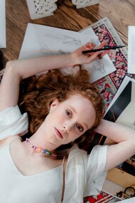 A fashion designer in a white blouse lies on a carpet, working on her sketches
