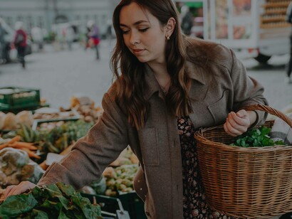 A woman food shopping at the farmer's market carrying a basket with some freshly cut produce