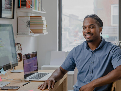 A man sits at a desk in a home office, talking on the phone