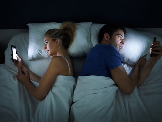 A husband and wife lie back-to-back in bed, each absorbed in scrolling through their phone
