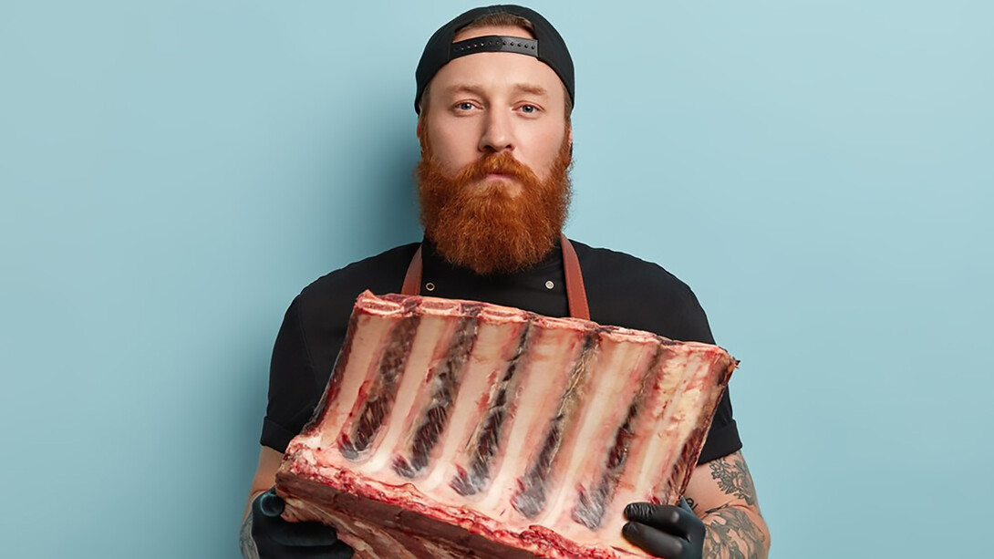 A man with a ginger beard, wearing an apron and gloves, holding high-tech food meat
