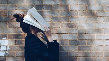 A schoolgirl stands against a rustic red brick wall, holding a book up to cover her face, reflecting student burnout