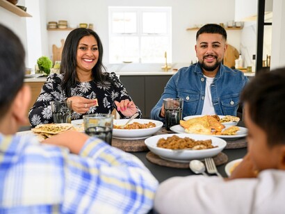 A three-generation Pakistani family shares a meal while a woman prepares food for the gathering