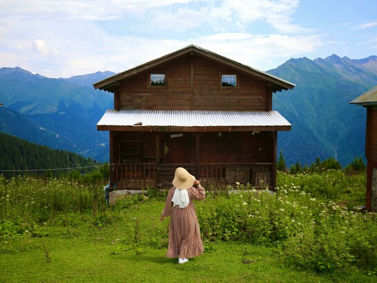 A woman wearing a hat walks toward a rustic wooden cottage surrounded by breathtaking mountain scenery and peaceful nature