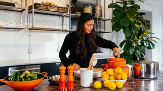 A woman stands gracefully in a kitchen, surrounded by an array of vibrant fruits, while elegantly holding the lid of a pot