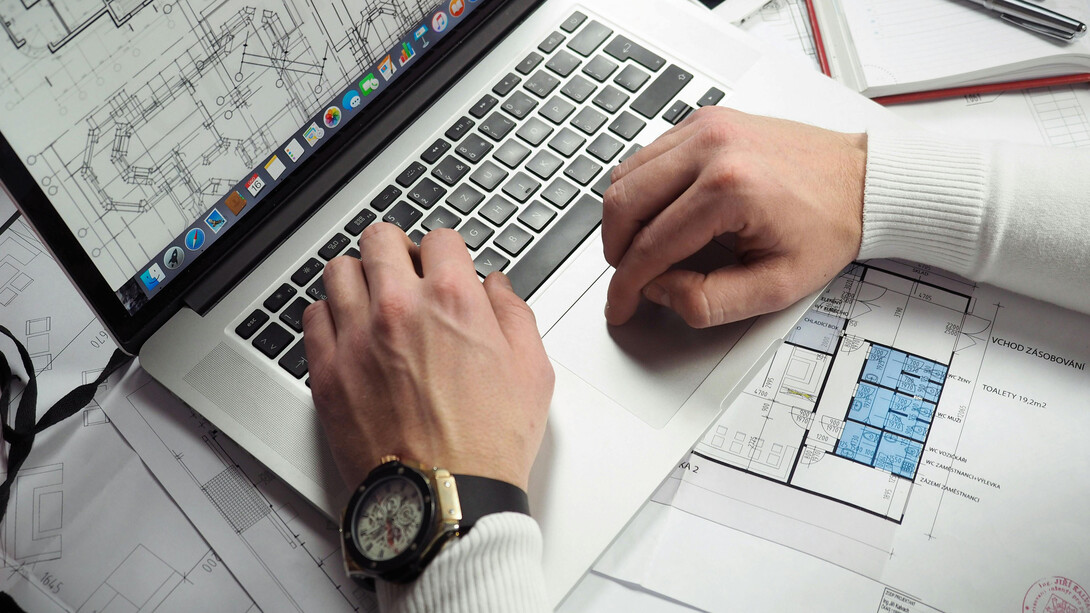The hands of a man sit on a laptop as he works on very detailed blueprints 