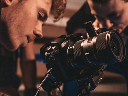 A man in a black shirt is holding a black DSLR camera on a film set, surrounded by Hollywood film crew members
