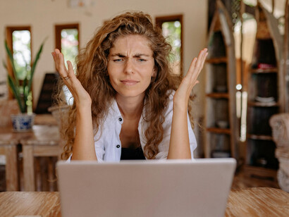 A woman in a white button-up shirt works on her laptop, her face marked by frustration