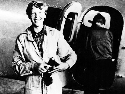 American aviator/pilot Amelia Earhart standing by her Lockheed Electra, dressed in overalls, with Fred Noonan getting into the plane in the background