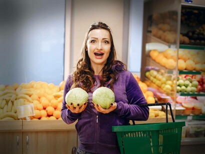 Una donna con un'espressione divertita tiene due cavoli verdi all'altezza del petto, in un reparto di frutta e verdura di un supermercato