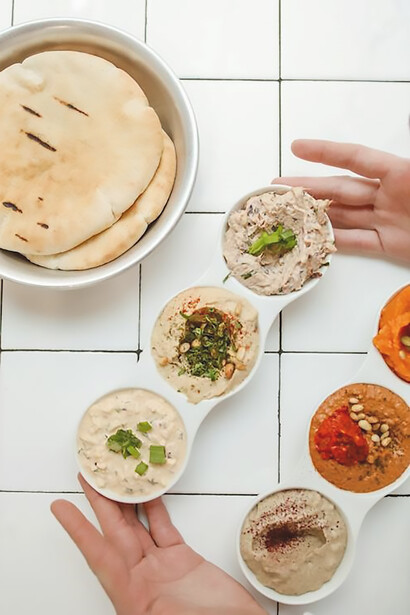 Upon a pristine white surface, a tempting selection of hummus varieties is tastefully presented, each one a visual symphony of flavors waiting to be savored