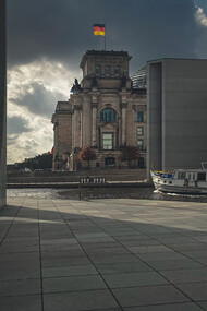 An elderly man walks along a gray concrete path beside a brown building in Berlin, Germany