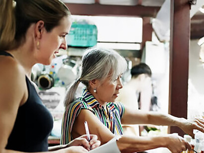 A senior Thai woman guides tourists through the process of cooking authentic Thai cuisine in a traditional kitchen setting