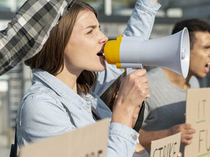 A woman passionately speaks through a megaphone during a public demonstration, surrounded by others holding protest signs, capturing the essence of active civic engagement and underscoring the importance of political involvement in shaping society and advocating for change