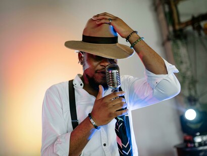 A man wearing a hat sings on stage with a band during a high-energy concert, featuring drums and a classic style