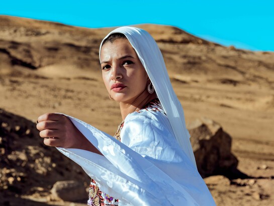 A woman wearing flowing white traditional attire against a desert landscape, representing cultural identity shaped by heritage and place