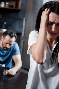 Depressed woman sitting in distress as her husband drinks alcohol in the background
