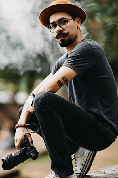 A man with a mustache, glasses, and a hat, dressed in a plain t-shirt and sneakers, posing confidently
