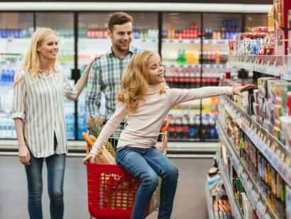 A happy young family with a child sits on a shopping cart in a supermarket aisle, surrounded by food items